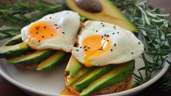 Two Toasts With Avocado And Fried Egg Spreading Yolk, Rosemary.